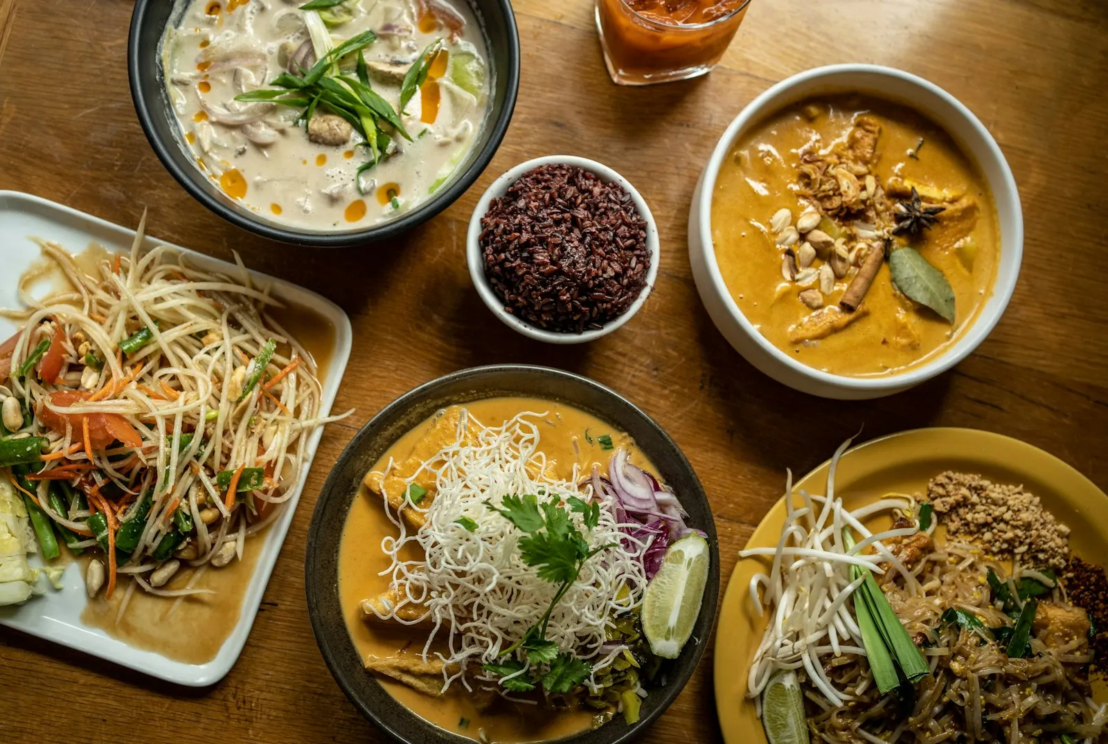 Wooden table covered with colorful plates of Thai dishes, curries, noodles and rice