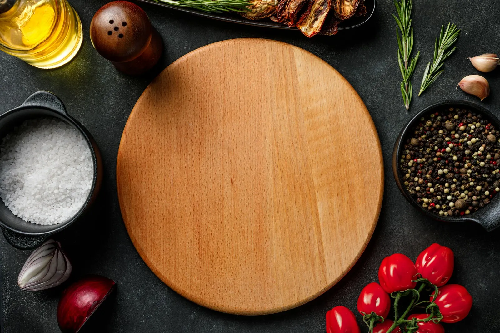 Wooden cutting board surrounded by fresh tomatoes, garlic, herbs, oil and spices on a dark table